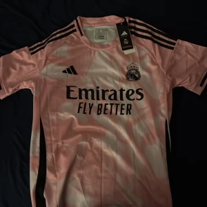 Rosa Real Madrid fotbollströja Adidas - Säljer en rosa Real Madrid fotbollströja från Adidas med svarta detaljer och klubbmärke på bröstet. Tröjan har korta ärmar, rund hals och är tillverkad i ett lätt, glansigt funktionsmaterial. Klassiska Adidas-ränder på axlarna och tryckt sponsorlogga framtill.
