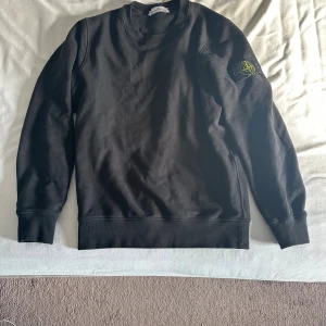 Svart sweatshirt från Stone Island - Svart långärmad sweatshirt från Stone Island med klassisk rund hals och ikonisk logotyp-patch på vänster ärm. Tröjan är i mjuk bomull och har ribbade muddar vid ärmslut och nederkant. Perfekt för en stilren och avslappnad look.