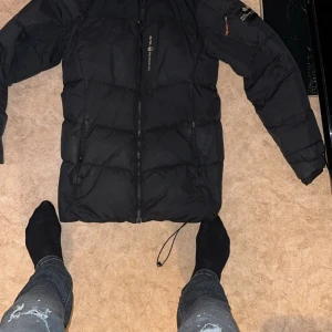 Svart pufferjacka Sail Racing - Svart pufferjacka från Sail Racing med huva och dragkedja framtill. Jackan har långa ärmar, justerbar dragsko nedtill och diskret logga på bröstet. Perfekt för kalla dagar och har en stilren look som funkar till jeans och sneakers.