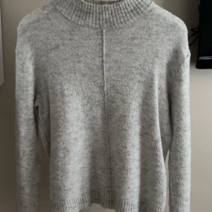 Grå stickad tröja från KappAhl - Mysig grå stickad tröja från KappAhl med hög ribbad krage och synlig söm framtill. Tröjan har lång ärm och ribbade muddar vid ärmkanten.