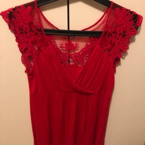 Röd klänning med spetsdetaljer - Supersnygg röd klänning med omlott framtill och vackra spetsdetaljer över axlar och rygg. Klänningen har en feminin v-ringning och är ärmlös med transparent spets upptill. Perfekt för dig som vill sticka ut med en färgstark look.