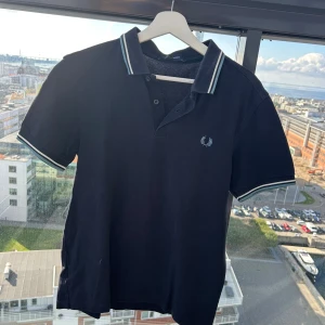 Svart pikétröja från Fred Perry - Klassisk svart pikétröja från Fred Perry med vit och ljusblå rand på krage och ärmslut. Tröjan har korta ärmar, knäppning framtill och den ikoniska Fred Perry-loggan broderad på bröstet. Perfekt för en stilren och sportig look.