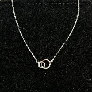Halsband med dubbla ringar i silver - Stilrent halsband i äkta silver med två sammanlänkade ringar, varav en är dekorerad med små glittrande stenar. Tunn kedja med justerbar längd (41-46cm) och diskret lås. Perfekt för dig som gillar minimalistiska smycken med en twist. Från glitter helt oanvänt nypris 300kr