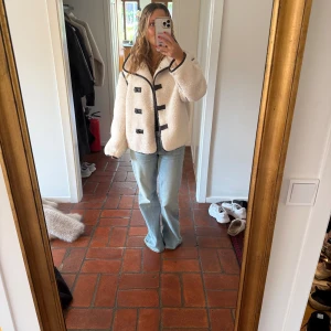 Teddyjacka  - Säljer en beige teddyjacka med svarta kanter och snygga knäppningar framtill. Jackan har en loose passform och är perfekt för dig som gillar en mysig och trendig stil. Fodrad och med stora krage för extra vibe.