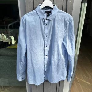 Ljusblå slim fit skjorta från H&M - Ljusblå skjorta från H&M i premium bomull, slim fit-modell. Skjortan har svarta knappar och snyggt mönstrat tyg på insidan av manschetterna och kragen. Perfekt för dig som gillar stilrena detaljer och en fräsch look.