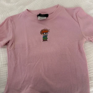 Rosa Rugrats t-shirt från Bershka - Säljer en ribbad rosa t-shirt från Bershka med broderad Rugrats-figur på bröstet. T-shirten har korta ärmar och rund halsringning. Perfekt för dig som gillar retro och tecknade serier!