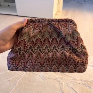 Becksöndergaard väska  - Säljer en unik kuvertväska från Becksöndergaard med broderat mönster i rosa, brunt och beige. Väskan har en mjuk form, innerfack med dragkedja och är tillverkad i textilmaterial med snyggt detaljerad design. Perfekt för att lyfta din outfit med lite extra färg och stil.