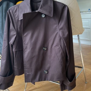 Brun kort trenchcoat med knappar - Kort brun trenchcoat med stora knappar framtill och på ärmsluten. Jackan har klassisk krage, två snedställda fickor och en snygg detalj med överlappande tyg på ryggen. Den har en liten ”clasp” vid halsen som man kan knäppa och overall så SJUKT snygg. Passar prefekt till hösten eller våren!!! Använd bara två gånger men helt underbar jacka!