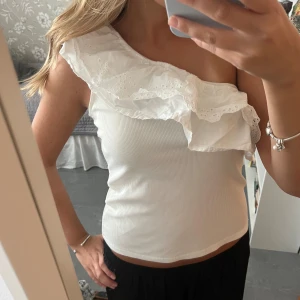 Vit oneshoulder topp med volang - Supersöt vit topp med oneshoulder och stor volangdetalj över axeln. Toppen är ribbad och har en romantisk känsla med broderad spets på volangen. Perfekt för dig som vill sticka ut lite extra i sommarvärmen.
