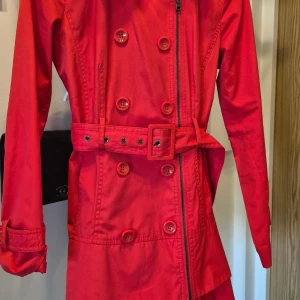 Röd trenchcoat från Guess XS - Snygg röd trenchcoat från Guess i storlek XS. Jackan har dragkedja, kapparna endast dekor kan ej knäppas, bälte i midjan och långa ärmar. Tillverkad i en mix av bomull och polyester. Perfekt för dig som vill sticka ut med en färgstark och klassisk look. 