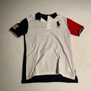 Pikétröja Polo Ralph Lauren - Snygg pikétröja från Polo Ralph Lauren med färgblock i vitt, marinblått och rött. Stor broderad logga på bröstet, stor siffra 3 på ryggen och amerikansk flagga på ärmen. Klassisk krage och korta ärmar. Perfekt för en sportig och stilren look.