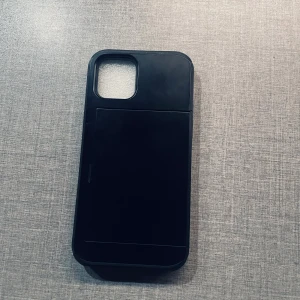 Spigen iPhone 12 Wallet Case - Robust svart Spigen plånboksfodral för iPhone 12 med kortfack. Fodralet är i mycket gott skick utan synligt slitage. Ger extra skydd och har plats för kreditkort eller ID. Perfekt för dig som vill ha både säkerhet och funktion i ett stilrent skal.