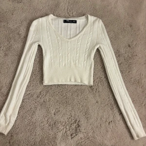 Långärmad magtröja från newyorker - Säljer en vit croppad stickad tröja från FB Sister. Tröjan har v-ringning, ribbade muddar och ett snyggt kabelstickat mönster. Perfekt för lager-på-lager och passar till både jeans och kjol. Långärmad och riktigt skön att ha på sig.