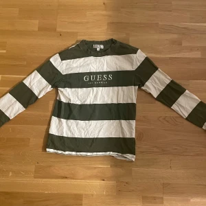 Randig långärmad tröja från Guess - Säljer en långärmad tröja från Guess med breda gröna och vita ränder och tryckt logga på bröstet. Tröjan har en klassisk passform och är gjord i mjuk bomull, perfekt för en chill och stilren look.