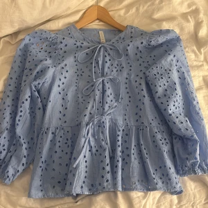 Blå broderad blus med knyt - Supersöt ljusblå blus från H&M med broderade blommor och hålmönster. Blusen har tre knytband framtill, volang nertill och puffiga långärmar med resår vid ärmslut. Perfekt för vår och sommar när du vill ha något luftigt och unikt.