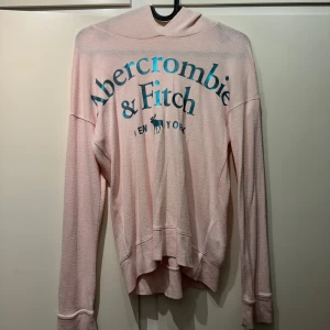 Ljusrosa hoodie från Abercrombie & Fitch - Supermjuk ljusrosa hoodie från Abercrombie & Fitch med stort blått tryck framtill. Tröjan har långa ärmar, huva och avslappnad passform. Perfekt för chill dagar och enkel att matcha med jeans eller leggings. STOR I STORLEKEN PASSAR xs och s Skriv för mer bilder och frågor så svarar jag så fort jag kan pris går altid att diskuteras💕💕