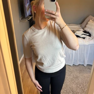 Beige stickad kortärmad topp - Säljer en stilren beige stickad topp med rund hals och korta ärmar. Toppen har en mjuk och följsam passform med ribbade muddar vid ärmslut och nederkant. Perfekt att matcha med både jeans och kjol för en enkel och snygg look.