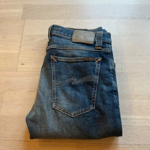 Mörkblå jeans från Nudie Jeans - Säljer ett par mörkblå jeans från Nudie Jeans med klassisk femficksdesign och snygga slitningar. Jeansen har raka ben och kontrastsömmar, tillverkade i mjukt denimtyg. Perfekta för dig som gillar en stilren och tidlös look. W31, L34 