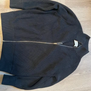 Svart stickad dubbel zipjacka Maison Margiela - Svart stickad zipjacka från Maison Margiela med ribbad struktur, klassisk krage och hel dragkedja framtill. Ikoniska vita stygn i nacken. Jackan har lång ärm och en clean, minimalistisk look som passar perfekt till streetwear eller lager-på-lager. Längd: 65cm Bröstvidd: 52 Armlängd: 60