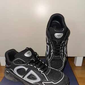 Svarta Dior B30 sneakers - Säljer ett par svarta Dior B30 sneakers med tydliga vita detaljer och reflekterande inslag. Skorna har en chunky sula, mesh och syntetmaterial samt stora CD-loggor på sidorna. Perfekta för dig som vill sticka ut med en lyxig och sportig vibe.