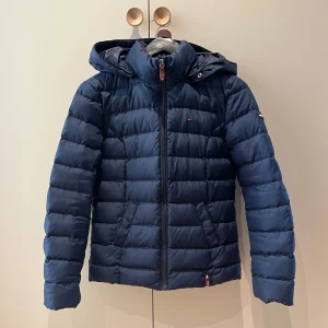 Blå pufferjacka från Tommy Hilfiger - Snygg marinblå pufferjacka från Tommy Hilfiger med huva och dragkedja framtill. Jackan har quiltad design, två fickor och diskret logga på bröstet. Perfekt för kyliga dagar och har en modern, normal passform.