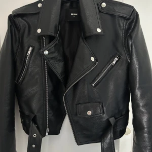 Svart skinnjacka från Bik Bok - Snygg svart skinnjacka från Bik Bok med klassisk bikerstil. Jackan har silvriga dragkedjor, knappar och ett brett bälte i midjan. Croppad passform, långa ärmar med dragkedjor och flera fickor framtill. Perfekt för dig som gillar edgy och stilren look. Nästintill oanvänd. 