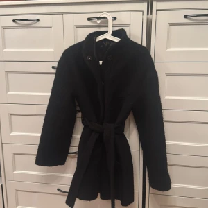 Svart teddyjacka från & Other Stories - Säljer en svart teddyjacka från & Other Stories med hög krage och knappar framtill. Jackan har bälte i midjan för snygg passform och är helfodrad. Perfekt för dig som vill ha en stilren och mysig jacka med trendig look.