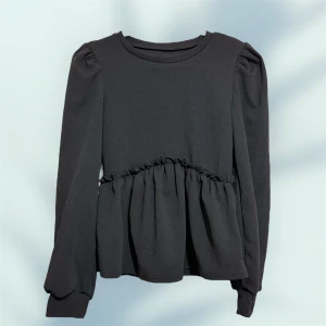 Svart långärmad blus med volang - Svart långärmad blus med rund halsringning och volangdetalj under bysten. Puffade axlar och breda muddar vid ärmslut ger en trendig siluett. Perfekt för dig som gillar stilrena och feminina plagg.