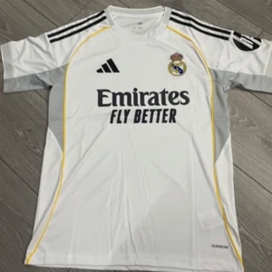 Real Madrid matchtröja Adidas S - Vit Real Madrid fotbollströja från Adidas i slim fit. Klassisk design med svarta ränder på axlarna, gula detaljer längs sidorna och klubbmärke på bröstet. Tillverkad i lätt och ventilerande material, perfekt för match eller träning.