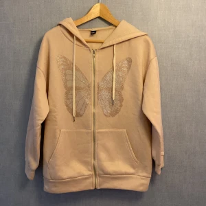Beige hoodie med fjäril från SHEIN - Superfin beige hoodie från SHEIN med glittrig fjäril på bröstet. Tröjan har dragkedja, huva med snören och stora fickor framtill. Perfekt för dig som gillar mysiga och snygga plagg med lite extra detalj.