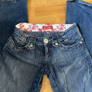 Blå lågmidjade jeans från VM Jeans med röda sömmar - Snygga blå jeans från VM Jeans med coola röda kontrastsömmar och detaljerade fickor. Insidan har ett unikt blommigt tyg i vitt och rosa. Klassisk femficksmodell och gylf med knapp. Perfekta för dig som gillar jeans med lite edge.