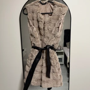 Beige pälsväst med bälte - Mysig beige pälsväst. Västen är ärmlös och har en fluffig, lyxig känsla som ger en cool look. Perfekt att slänga över en hoodie eller långärmad tröja för extra stilpoäng, kan även användas över en svart skinnjacka. 