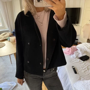 Svart kort kappa med knappar - Säljer en svart, kort kappa med dubbelknäppning och rak passform. Jackan har långa ärmar och en stilren look som passar perfekt till jeans eller kjol. Materialet känns mjukt och har en klassisk känsla. Enkel och snygg att slänga på sig när det är kyligt ute. Från Gina tricot 