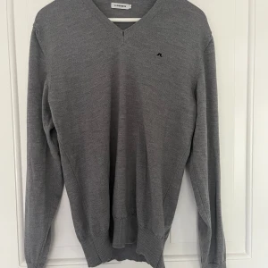 V-ringad J.Lindeberg tröja  - Snygg grå v-ringad tröja från J.Lindeberg med liten broderad logga på bröstet. Tröjan är stickad i ett tunt material och har långa ärmar samt ribbade muddar vid ärmslut och nederkant. Perfekt för lager-på-lager under hösten och vintern. Ett stygn i v-ringningen har släppt (se bild 3). Sitter som M. Hör av dig vid funderingar 👍