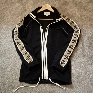 Gucci tracksuit - Säljer en svart ziptröja från Gucci med vita detaljer och breda logoband längs ärmarna. Plagget är i väldigt gott skick. Tröjan har hel dragkedja framtill, snörning nertill och är tillverkad i 100% bomull. Snygg och sportig look med ikoniska GG-loggor på ärmarna. Plagget är i storlek M men passar S också (Pris går att diskutera)