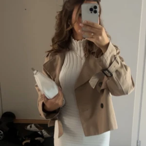 Beige croppad trenchcoat - Snygg croppad trenchcoat i beige med dubbelknäppning och bältdetaljer vid ärmsluten. Jackan har klassisk krage och svarta knappar, perfekt för dig som vill ha en trendig och stilren look.