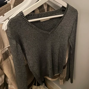 Glittrig mörkgrå stickad tröja - Snygg mörkgrå stickad tröja med glittriga detaljer och v-ringad hals. Tröjan har långa ärmar och ribbade muddar vid hals, ärmslut och nederkant. Perfekt för dig som vill ha lite extra shimmer i outfiten.