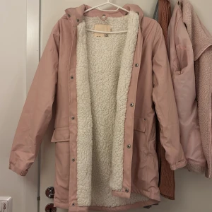 Rosa parkajacka med teddyfoder från 157 - Mysig rosa parkajacka från 157 med huva och vitt teddyfoder på insidan. Jackan har stora fickor framtill, tryckknappar och reflexdetalj baktill. Perfekt för kalla dagar när du vill hålla dig varm och snygg.