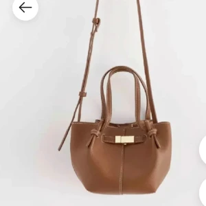 Brun väska Lindex - Snygg brun axelväska i skinnimitation med justerbar axelrem och två handtag. Väskan har dekorativ gulddetalj framtill och kontrastsömmar som ger en stilren look. Perfekt storlek för det viktigaste och passar till många outfits.
