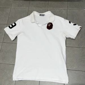Vit  BAPE Polo - Vit polo från A Bathing Ape med klassisk BAPE-logga broderad på bröstet och svart tryck med siffror på båda ärmarna. Polon har krage och är perfekt för dig som gillar streetwear och vill sticka ut med en ikonisk look.