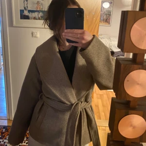 Beige omlott kappa med bälte - Säljer denna supersnygga kappa då den inte passar så bra längre. Den är lite mindre i storleken men går att justera med bandet runt om. Super skön nu till hösten! Jackan är använd ett fåtal gånger 🤍 hör av er för frågor eller fler bilder! 