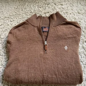 Brun halvziptröja från Morris - Snygg brun stickad tröja från Morris med halv dragkedja och broderad logga på bröstet. Tröjan har ribbade muddar och hög krage, perfekt för lager-på-lager. Skön och stilren modell som passar till jeans eller chinos.