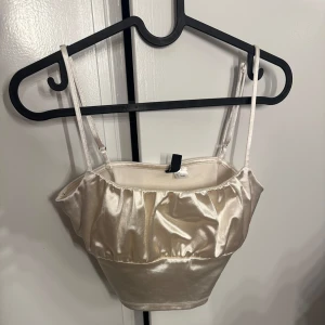 Champagnefärgad satin bandeau från H&M - Säljer en glansig champagnefärgad bandeau topp från H&M Divided. Toppen har smala justerbara axelband och en volangdetalj framtill. Materialet är satinliknande och ger en lyxig känsla. Perfekt för dig som vill sticka ut med en elegant och trendig look.