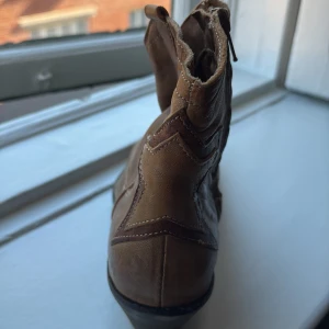 Bruna cowboy boots i skinn - Säljer ett par snygga bruna cowboy boots i skinn med klassiska broderade detaljer på tån och dekorativa sömmar. Bootsens dragkedja på sidan gör dem enkla att ta på och av. Perfekta för dig som vill ha western-vibe i din stil.Köpta i st tropez