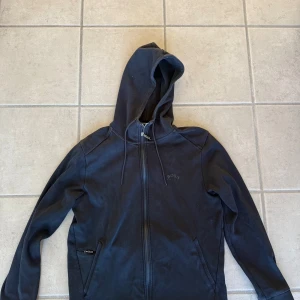 Svart hoodie från Hugo Boss med dragkedja - Svart hoodie från Hugo Boss med dragkedja framtill. Tröjan har diskret Boss-logga på bröstet och två fickor med dragkedja. Tillverkad i mjuk bomullsblandning för extra komfort.