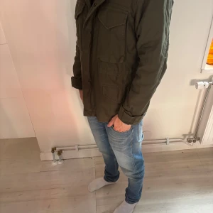 Grön parkajacka från Superdry - Säljer en mörkgrön parkajacka från Superdry med flera stora fickor och quiltad insida. Jackan har långärmade ärmar, knappar och dragkedja framtill samt fleecefoder för extra värme. Perfekt för dig som gillar streetwear och funktionella detaljer.