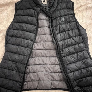 Svart dunväst  - Snygg svart dunväst med quiltad design och dragkedja framtill. Västen har en hög krage och är fodrad i grått på insidan. Perfekt att slänga över en hoodie eller tröja för extra värme. I stlk Xs - S