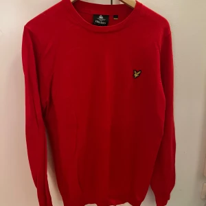 Röd stickad tröja från Lyle & Scott - Riktigt snygg röd stickad tröja från Lyle & Scott med klassisk rund hals och deras ikoniska gula fågel-logga på bröstet. Tröjan har långa ärmar och ribbade muddar vid ärmslut och nederkant. Perfekt för dig som vill ha en stilren och färgstark look.