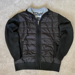 Hybrid Knit Cardigan - Svart kofta med quiltad framsida och stickade ärmar och rygg. Insidan har ett grått och svart camouflagemönstrat fleecefoder. Dragkedja framtill och ribbade muddar vid ärmslut och nederkant. Perfekt för kyligare dagar.