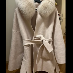 Beige kappa med päls och bälte - Superfin beige kappa med stor fluffig päls runt kragen och knytskärp i midjan. Jackan har stora fickor framtill och en avslappnad, loose passform. Perfekt för dig som vill ha en mysig och trendig look under kyliga dagar.